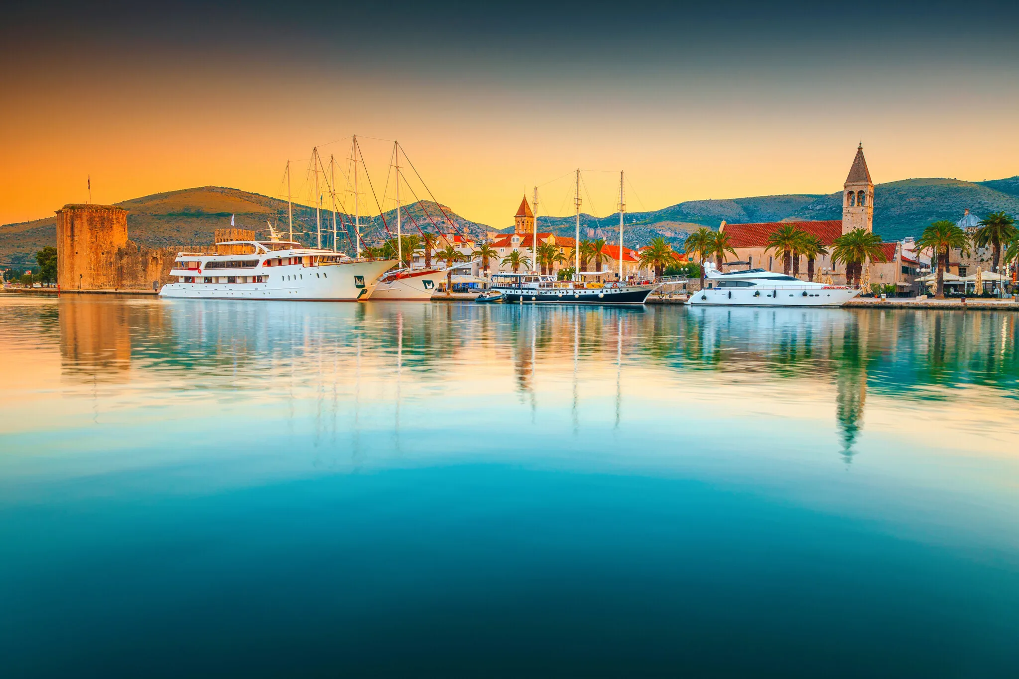 Colorful sunrise at harbor of Medieval and historic old town, Trogir, Croatia. Trogir is a historic town and harbour on the Adriatic coast in Split-Dalmatia county. Visible are many sailboats, yachts, Fortress Kamerlengo, palm trees and beautiful morning reflection in the water.