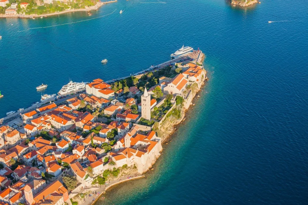 Tip of Rab Island seen from above, with old town with red roofs and a chuch spire, and two large motor yachts moored along the shore.