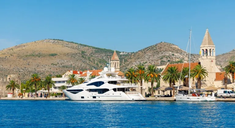 A white superyacht moored stern-to on the Trogir waterfront in Croatia, with the cathedral of St Lawrence and palm trees behind it