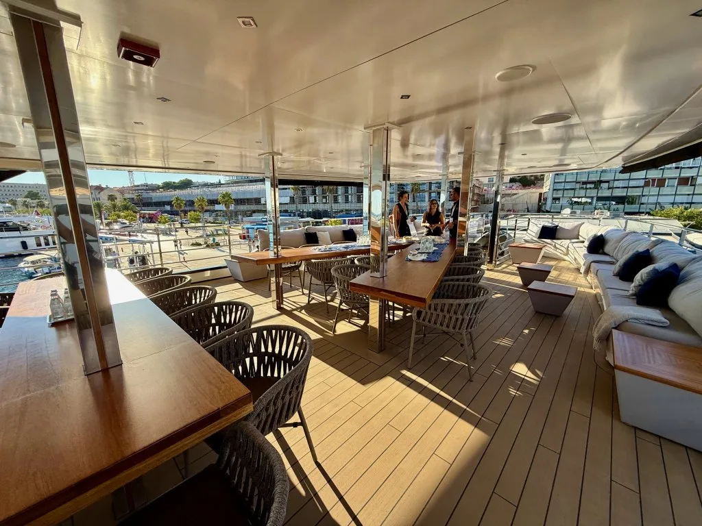 The shaded bridge deck of motor yacht ARGO with its long dining table and lounge, Split harbor behind, September 2026.
