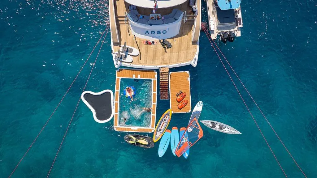 Motor yacht ARGO seen from above at anchor with the inflatable sea pool, floating platforms, paddleboards and chase boat deployed, from the yacht's own photography.