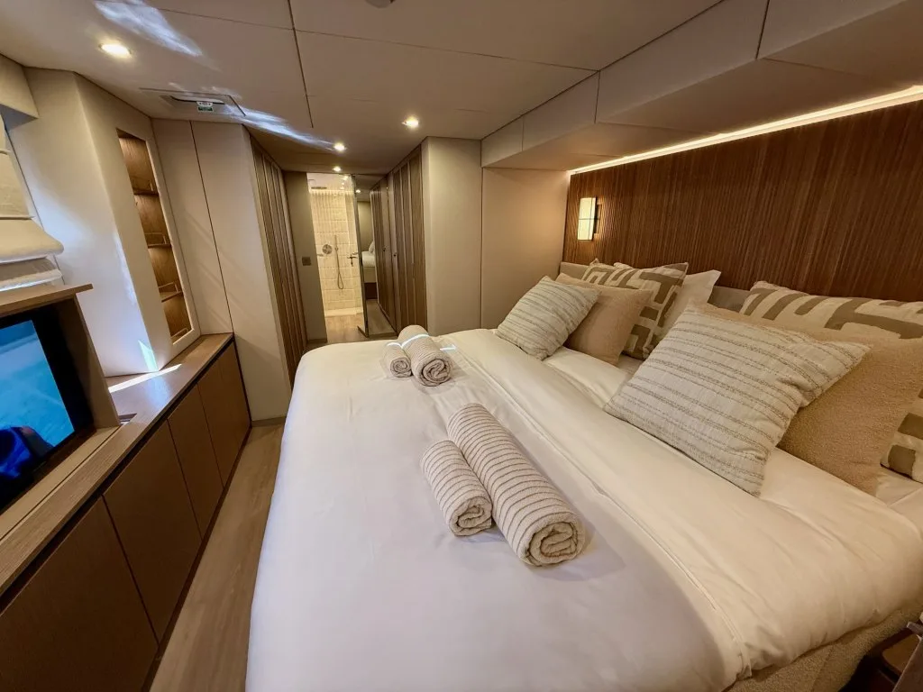 The master cabin of catamaran MIDORI with a wide bed under a slatted wood headboard and a window to the sea, September 2026.