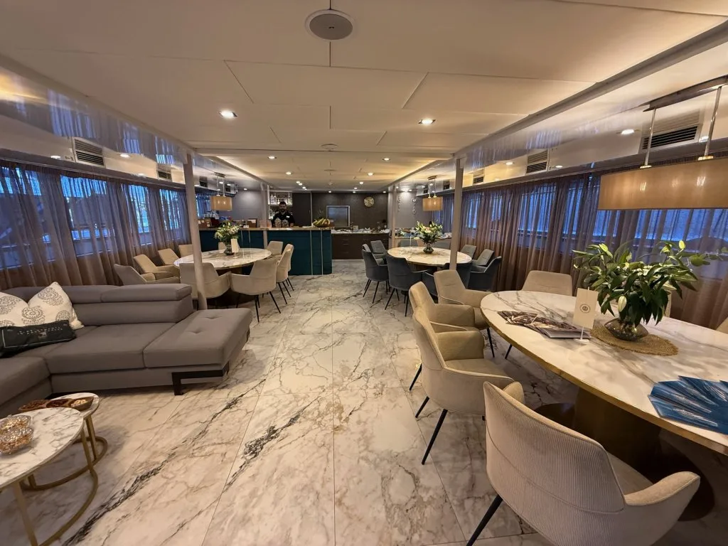 The main saloon of motor yacht OHANA with marble floors, a grey lounge and several round dining tables, September 2026.
