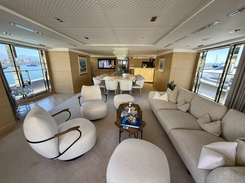 The main saloon of motor yacht RENATA with curved cream armchairs, a long sofa and the dining area beyond, September 2026.