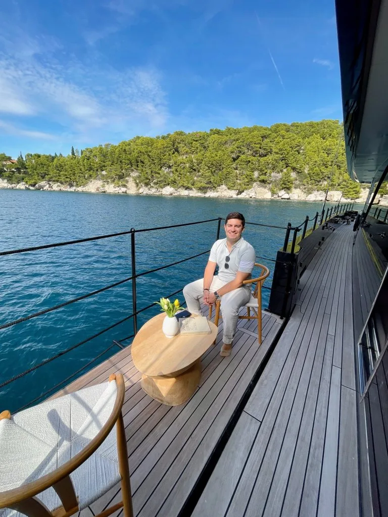 A DMA team member seated on the fold-down balcony of motorsailer REPOSADO, sea and coast behind, September 2025.