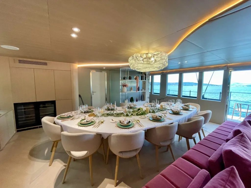 The main saloon of motorsailer REPOSADO with the oval dining table laid in green and gold under a shell chandelier, burgundy sofa beside it, September 2025.