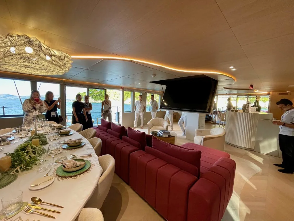 The main saloon of motorsailer REPOSADO with the burgundy lounge, dining table and screen, guests touring during the show, September 2025.